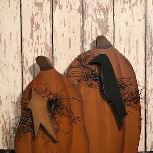 May include: Two wooden pumpkin-shaped cutouts with brown stained finishes and dark stems. The smaller pumpkin features a tan star cutout, while the larger one displays a black bird silhouette. Both are accented with dark brown dried raffia.