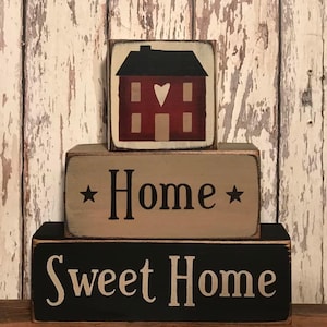 May include: A three-tiered wooden block set with a home decor theme. The top block features a red house with a black roof and a heart-shaped cutout. The middle block reads "Home" with star accents, and the bottom block says "Sweet Home."