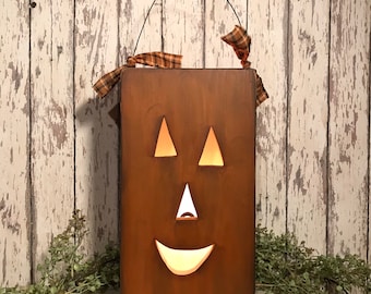 May include: A rectangular brown wooden jack-o'-lantern with two triangular eyes, a triangular nose, and a curved mouth cutout. The piece features a thin black wire handle at the top and is decorated with a brown and orange plaid fabric ribbon.