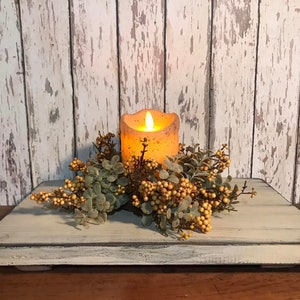 May include: A rustic, rectangular wooden tray with a distressed, off-white finish. It features a battery-operated candle with a flickering flame, surrounded by a decorative arrangement of green leaves and yellow berries. The background is a weathered, white wooden wall.