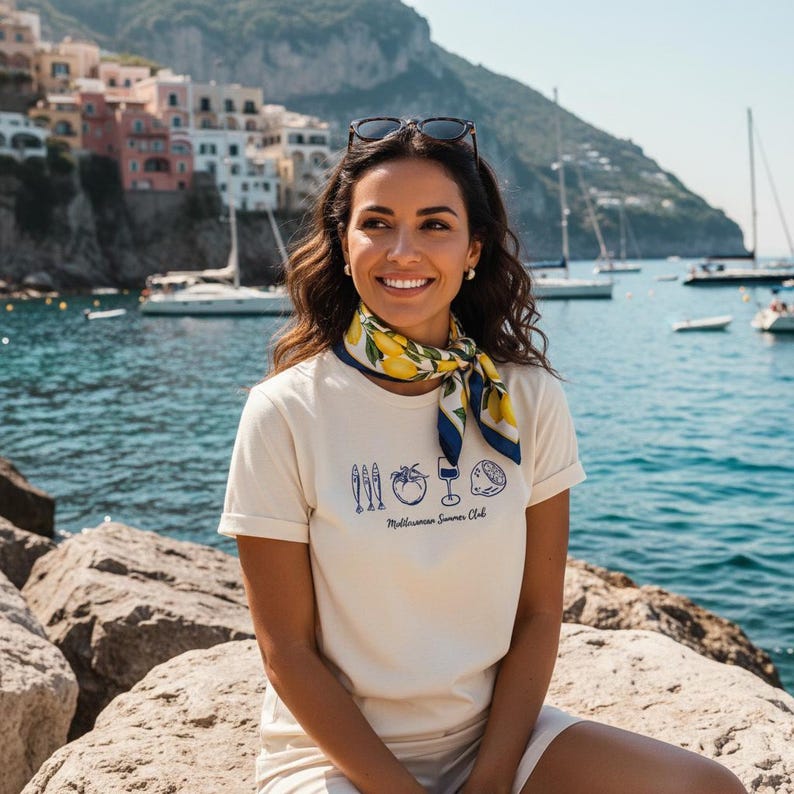 Puede incluir: Una camiseta de manga corta color crema con un estampado gr&aacute;fico azul de tres peces, un tomate, una copa de vino y un lim&oacute;n, con el texto Mediterranean Summer Club. Un pa&ntilde;uelo amarillo y azul con estampado de limones y gafas de sol oscuras.