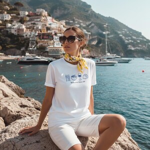 Puede incluir: Una camiseta blanca de manga corta con un estampado gr&aacute;fico azul de peces, un tomate, una copa de vino y un lim&oacute;n, con el texto "Mediterranean Summer Club" debajo. Se combina con un pa&ntilde;uelo amarillo con estampado de limones.