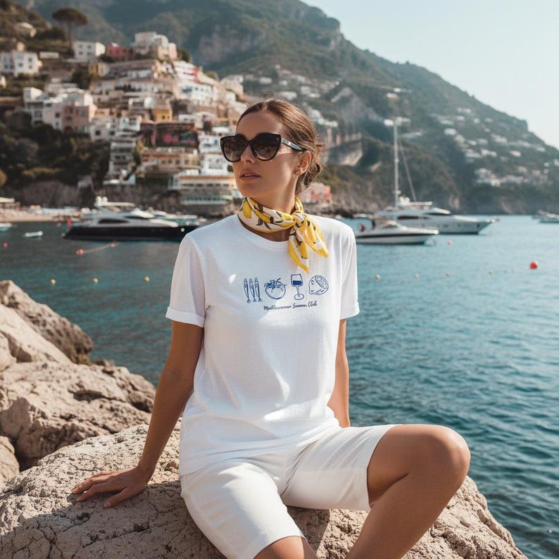 Puede incluir: Una camiseta blanca de manga corta con un estampado gr&aacute;fico azul de peces, un tomate, una copa de vino y un lim&oacute;n, con el texto "Mediterranean Summer Club" debajo. Se combina con un pa&ntilde;uelo amarillo con estampado de limones.