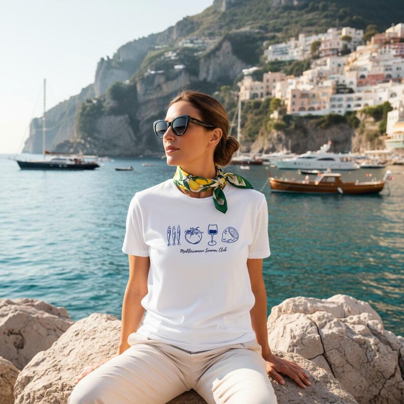 Puede incluir: Una camiseta blanca de manga corta con un estampado gr&aacute;fico azul de tres peces peque&ntilde;os, un tomate, una copa de vino y un lim&oacute;n, con el texto Mediterranean Summer Club debajo. Se lleva un pa&ntilde;uelo de cuello con estampado verde y amarillo.