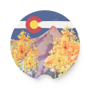 May include: A round coaster featuring a watercolor painting of a mountain range with yellow aspen trees in the foreground. The top of the coaster has a blue and white Colorado state flag with a red circle and a yellow star.