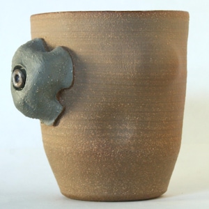 May include: A cylindrical ceramic mug with a matte, earthy brown finish and a textured surface. Attached to the side is a dark grey, rock-climbing hold shaped handle featuring a small, circular, metallic-looking bolt detail in the centre.