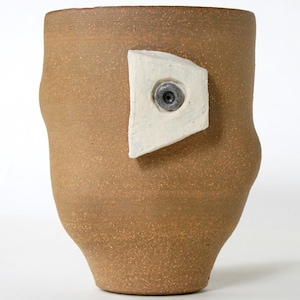May include: A brown ceramic mug with a white, geometric shape attached to the side. The shape has a small, silver-colored circle in the center.