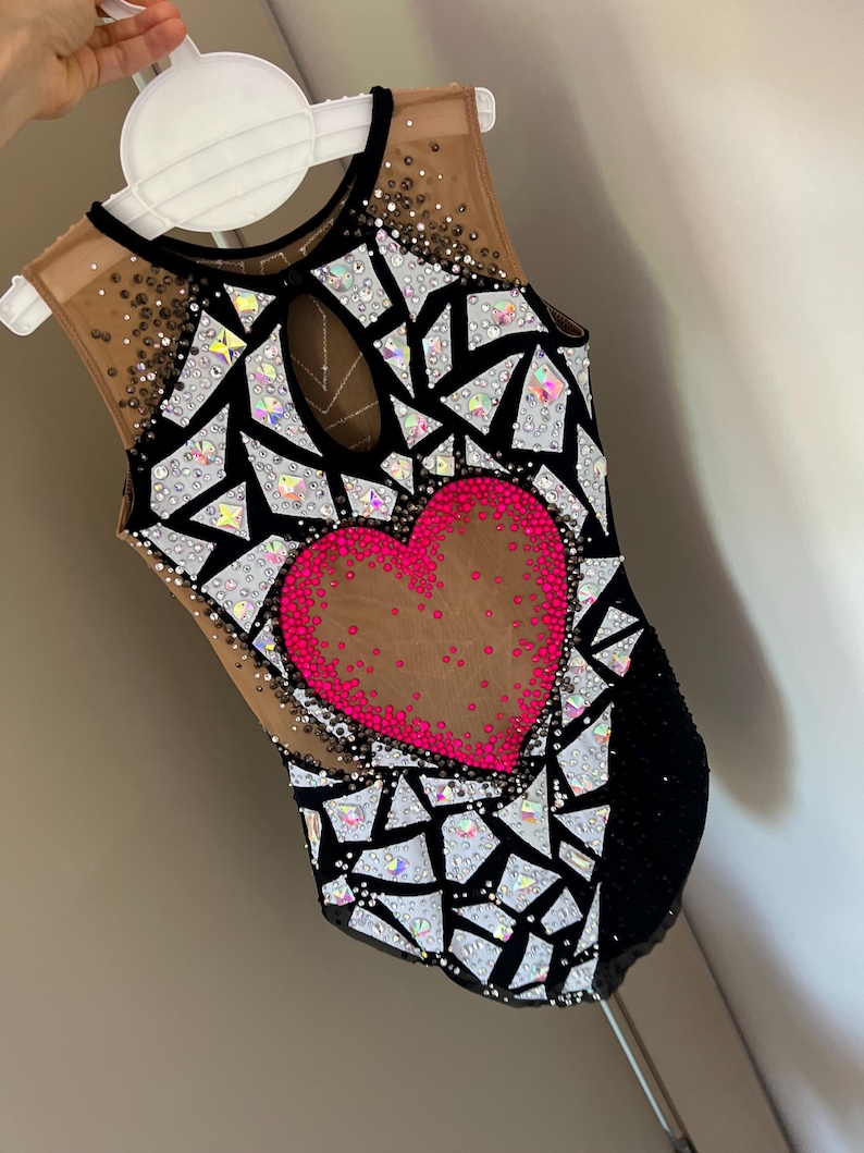 May include: A sleeveless leotard featuring a black and nude mesh base with a central hot pink heart motif. The garment is heavily embellished with geometric white and iridescent crystal shards, along with scattered small dark and clear rhinestones.
