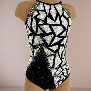 May include: A sleeveless leotard featuring a black base with a geometric pattern of white triangular shapes. The garment is heavily embellished with iridescent crystals and rhinestones, with sheer nude mesh fabric at the upper chest and shoulders.