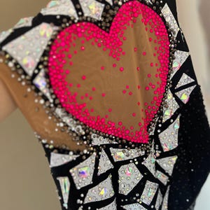 May include: A black leotard featuring a large central heart shape outlined in vibrant hot pink rhinestones on a nude mesh panel. The garment is heavily embellished with a mosaic pattern of white and iridescent crystals on black fabric.