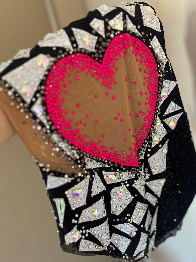 May include: A black leotard featuring a large central heart shape outlined in vibrant hot pink rhinestones on a nude mesh panel. The garment is heavily embellished with a mosaic pattern of white and iridescent crystals on black fabric.