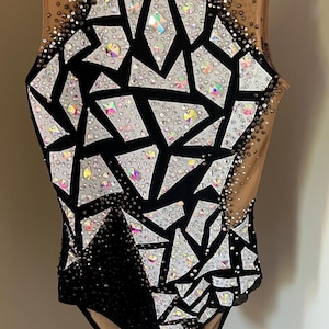 May include: A sleeveless leotard featuring a black and nude mesh base with a geometric pattern of white fabric shards. The garment is heavily embellished with iridescent and clear crystals of varying sizes, creating a sparkling effect across the front.