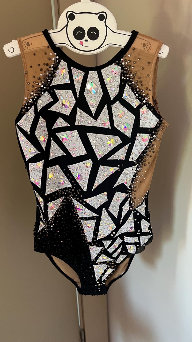 May include: A sleeveless leotard featuring a black and nude mesh base with a geometric pattern of white fabric shards. The garment is heavily embellished with iridescent and clear crystals of varying sizes, creating a sparkling effect across the front.