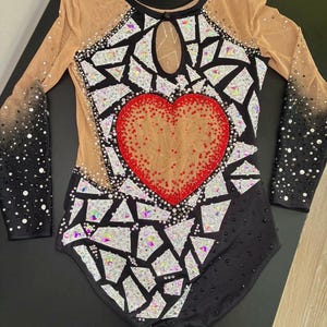 May include: A long-sleeved leotard featuring a black base with a central red heart motif. The garment is decorated with numerous iridescent, geometric, mirror-like shards and scattered crystals. It includes sheer beige mesh panels at the shoulders and chest, with a keyhole neckline and black sleeves that transition into a dense crystal pattern.