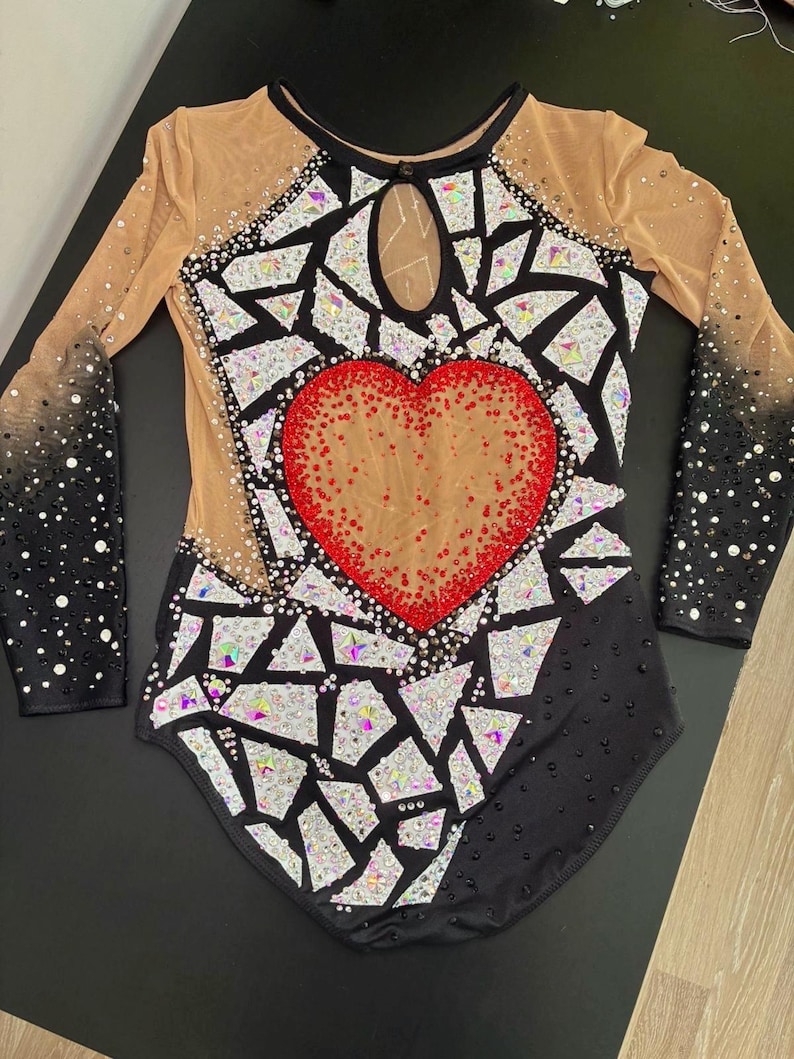 May include: A long-sleeved leotard featuring a black base with a central red heart motif. The garment is decorated with numerous iridescent, geometric, mirror-like shards and scattered crystals. It includes sheer beige mesh panels at the shoulders and chest, with a keyhole neckline and black sleeves that transition into a dense crystal pattern.