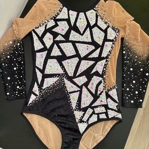 May include: A long-sleeved leotard featuring a black and nude mesh base with a geometric mosaic pattern of white and iridescent crystals. The sleeves transition from nude mesh to black fabric, heavily embellished with black and clear rhinestones.