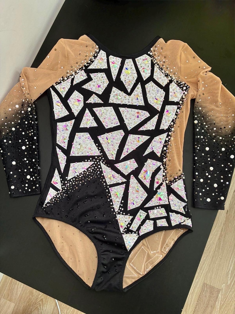 May include: A long-sleeved leotard featuring a black and nude mesh base with a geometric mosaic pattern of white and iridescent crystals. The sleeves transition from nude mesh to black fabric, heavily embellished with black and clear rhinestones.