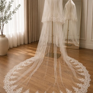 May include: A long, ivory-colored wedding veil featuring two tiers of sheer tulle fabric. The edges of the veil are adorned with intricate floral lace embroidery that extends along the entire hemline, creating a scalloped, decorative border.