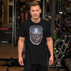 May include: Black short-sleeve t-shirt featuring a graphic design of a UFO hovering over a building. The design is framed within a skull-shaped outline. The shirt is worn by a person in a gym setting.