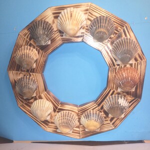 May include: A decorative wooden wreath with a geometric design, featuring a series of seashells. The shells are in various shades of beige, brown, and tan, set against a light blue background. The wooden frame has a burnt wood finish.
