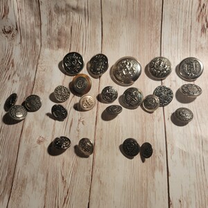 Könnte beinhalten: Eine Sammlung von silbernen und goldfarbenen Knöpfen mit verschiedenen Designs, darunter Wappen, Anker und geometrische Muster. Die Knöpfe sind auf einer hellbraunen Holzoberfläche angeordnet.