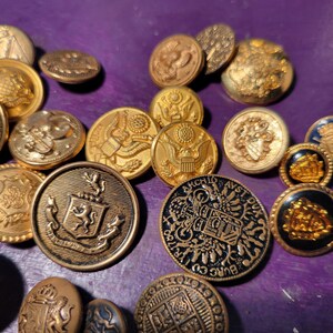 Könnte beinhalten: Eine Sammlung von Vintage-Knöpfen in Goldtönen mit verschiedenen Designs, darunter Wappen, Adler und florale Muster. Einige Knöpfe haben schwarze Emaille-Akzente.