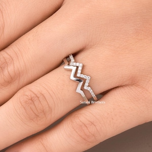 May include: A silver ring featuring a zigzag pattern, embellished with tiny, glittering stones. The ring is displayed on a finger, highlighting its detailed design and delicate features. The ring's design is contemporary and striking.