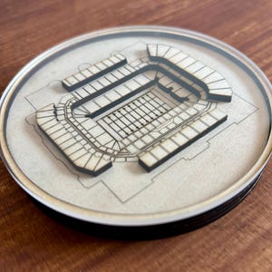 May include: A wooden coaster featuring a detailed laser-cut design of a football stadium. The coaster is circular and has a clear acrylic top.