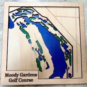 May include: A wooden map of the Moody Gardens Golf Course. The map is laser cut and painted with blue water and green grass. The map is framed in a light wood frame.