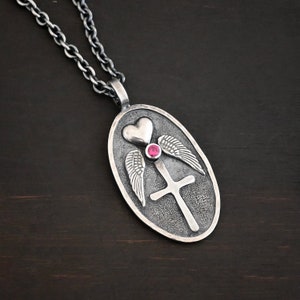 Puede incluir: Collar con colgante de plata en forma de cruz, corazón y alas. La cruz tiene una piedra preciosa roja en el centro. El colgante es de forma ovalada.