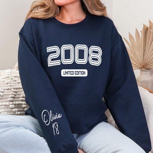 May include: Navy blue sweatshirt with the year "2008" in white, outlined text. Below the year is the text "LIMITED EDITION". The sleeve features the name "Olivia" and the number "18" in white script. The sweatshirt is a casual style.