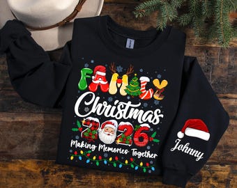 May include: A black long-sleeve sweatshirt featuring a colorful graphic that reads Family Christmas 2026 Making Memories Together. The design includes a Santa face, a Christmas tree, and holiday lights. The sleeve features a Santa hat and the name Johnny.