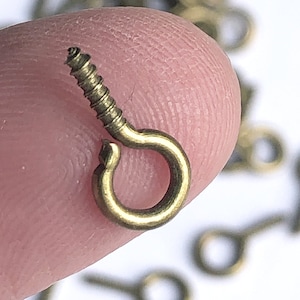 May include: A close-up of a gold-toned screw-in eye hook. The hook is small and has a simple design. It is perfect for hanging lightweight items.