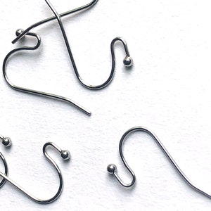 May include: Close-up of several silver-colored earring hooks. Each hook has a curved design with a small ball at the end of the curved section. The hooks are scattered on a white surface.
