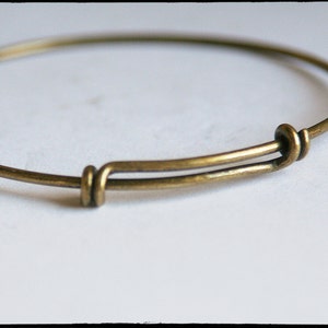 May include: Adjustable brass bangle bracelet. The bracelet features a simple, minimalist design with a sliding mechanism for size adjustment. The metal has a warm, antique brass finish, giving it a vintage look. The bracelet is shown against a white background.