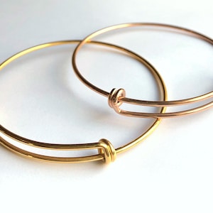 May include: Two adjustable bangle bracelets. One is gold-colored, the other is rose gold. Each bracelet has a simple, elegant design with a knot detail at the adjustable points. The bracelets are displayed on a white background.