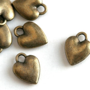 May include: A group of six antique gold heart-shaped charms with a loop at the top. The charms are scattered on a white surface.