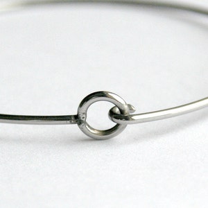 May include: A close-up of a silver-colored bangle bracelet. The bracelet features a simple, minimalist design with a circular clasp. The metal has a shiny, reflective surface. The background is a soft, neutral white.
