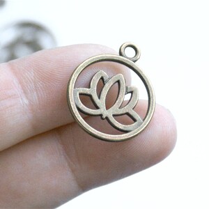 May include: A bronze-colored metal charm with a cutout design of a lotus flower. The charm has a loop at the top for attaching to a necklace or bracelet.