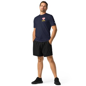 May include: Navy blue t-shirt with a pink heart graphic and the word "RUNNING" in yellow. The shirt is paired with black shorts and black athletic shoes. The person is standing with their hands in their pockets.