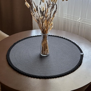 May include: A round, dark grey woven placemat with a thick, braided black border. Centred on the placemat is a clear glass vase containing a bouquet of dried lavender stems and light-coloured, fluffy, oval-shaped botanical seed heads.