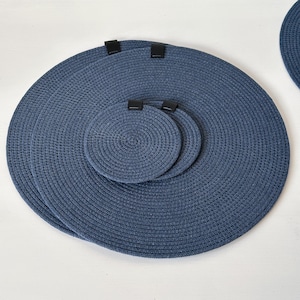 May include: A collection of navy blue circular placemats and coasters. The placemats and coasters are made from a woven material, featuring a spiral design. The placemats have black clips attached. The coasters are smaller and share the same design as the placemats.