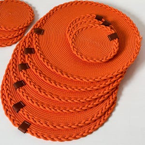 May include: A set of round orange placemats and matching coasters made from coiled rope. Each piece features a braided rope border and small brown leather tabs attached to the edges. The items are arranged in a stack on a flat surface.