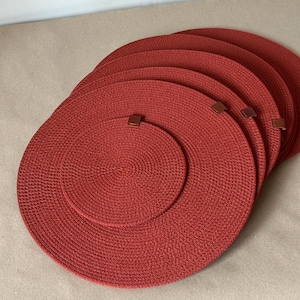 May include: A set of circular, rust-colored woven placemats featuring a dense, coiled rope texture. Each placemat is finished with a small, brown leather-like tab attached to the outer edge, providing a consistent design across the stack.