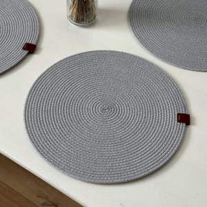 Peut inclure: Un ensemble de sets de table circulaires gris clair fabriqués à partir de corde enroulée. Chaque set présente un motif en spirale cohérent et une petite languette rectangulaire marron foncé en similicuir fixée sur le bord extérieur.