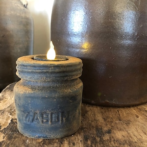May include: A rustic, dark blue mason jar candle holder with a lit candle inside. The candle holder is sitting on a wooden surface next to a brown ceramic jug.