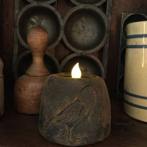 Könnte beinhalten: Ein dunkelbrauner Kerzenhalter aus Keramik mit einem geschnitzten Vogelmotiv. Der Kerzenhalter hat oben eine kleine, runde Öffnung und eine warme, gelbe Kerzenflamme im Inneren.