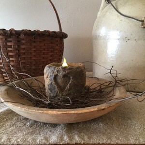 May include: A rustic, battery-operated candle with a carved heart design, emitting a warm, flickering light. The candle sits in a wooden bowl with dried twigs, alongside a woven basket and a stoneware jug. The scene evokes a cozy, farmhouse aesthetic.