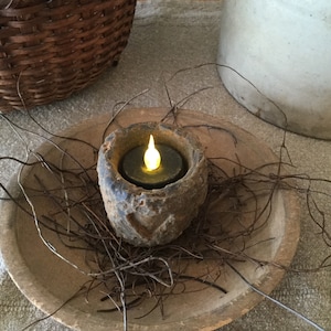 May include: A rustic, battery-operated candle in a weathered stone holder, emitting a warm, flickering light. The candle sits in a wooden bowl, surrounded by twigs, creating a cozy, natural aesthetic. A woven basket and a ceramic jug are in the background.