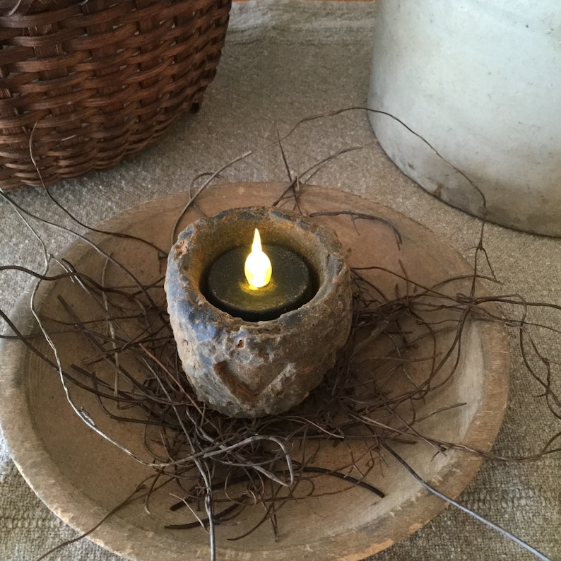 May include: A rustic, battery-operated candle in a weathered stone holder, emitting a warm, flickering light. The candle sits in a wooden bowl, surrounded by twigs, creating a cozy, natural aesthetic. A woven basket and a ceramic jug are in the background.